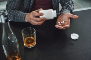 Man pouring medication tablets representing Sudafed into palm near glass of alcoholic beverage
