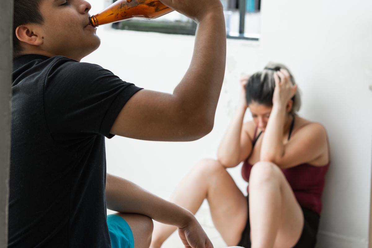Man drinking alcohol with woman holding her head and slumping against wall in background